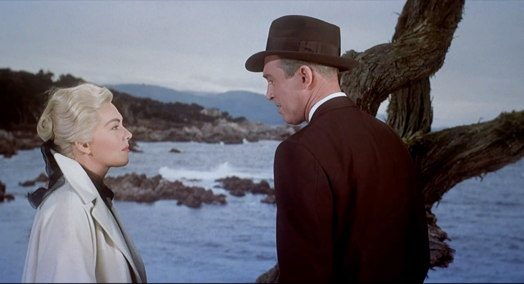James Stewart and Kim Novak stand in front of a rocky beach in fifties overcoats in this scene from Hitchcock's "Vertigo."
