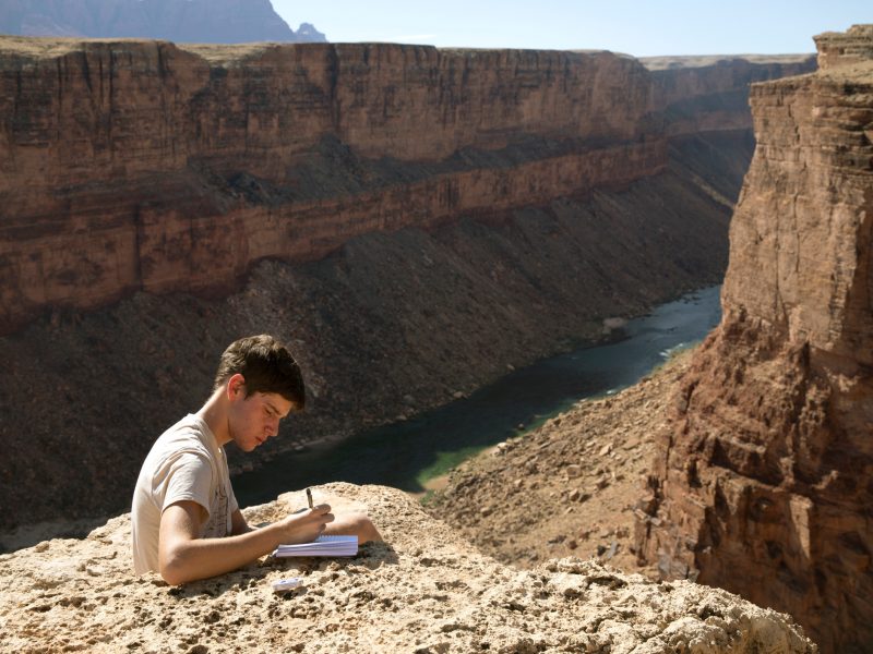 Student writes in a journal outside, with the Colorado River behind him.