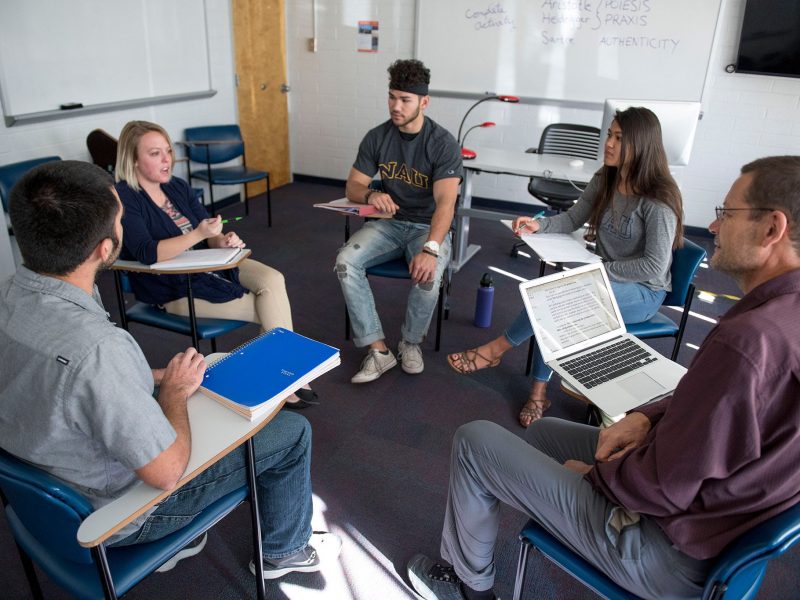 A group of students and professors sitting in desks in a circle talking to each other