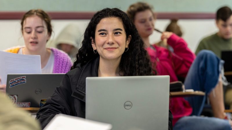 A girl sitting at a classroom desk with a laptop smiling