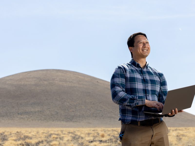 Faculty member outside smiling while holding a laptop.