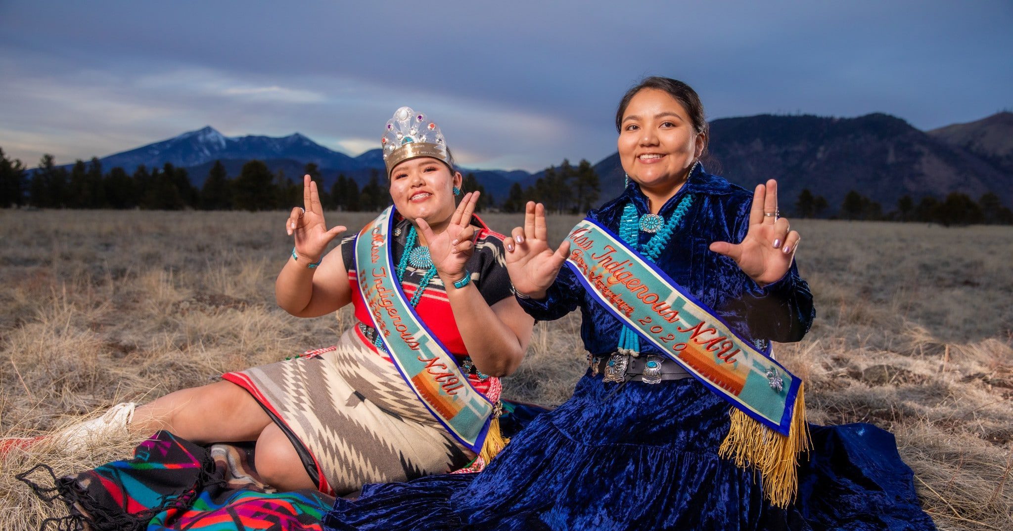 Dos mujeres en vestuario tradicional de Nativos Americanos sentadas en un prado enfrente de los San Francisco Peaks.