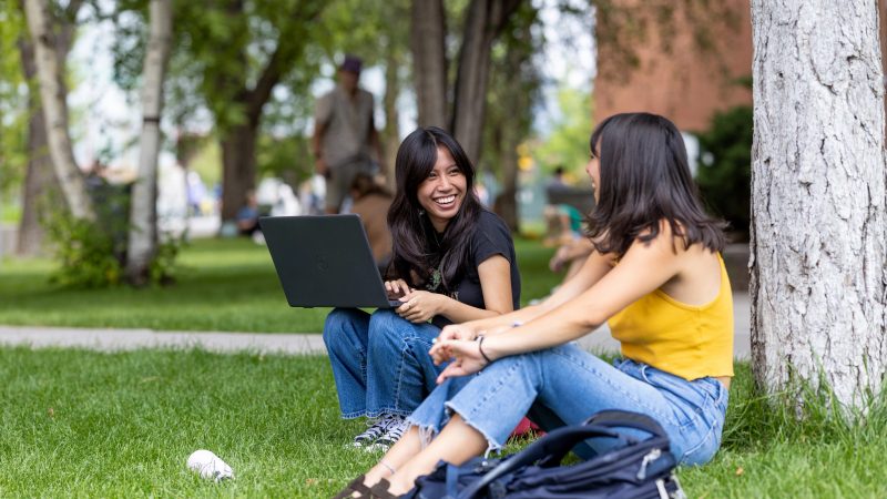 Dos estudiantes hablan mientras están sentados bajo del árbol en North Quad del campus de NAU.