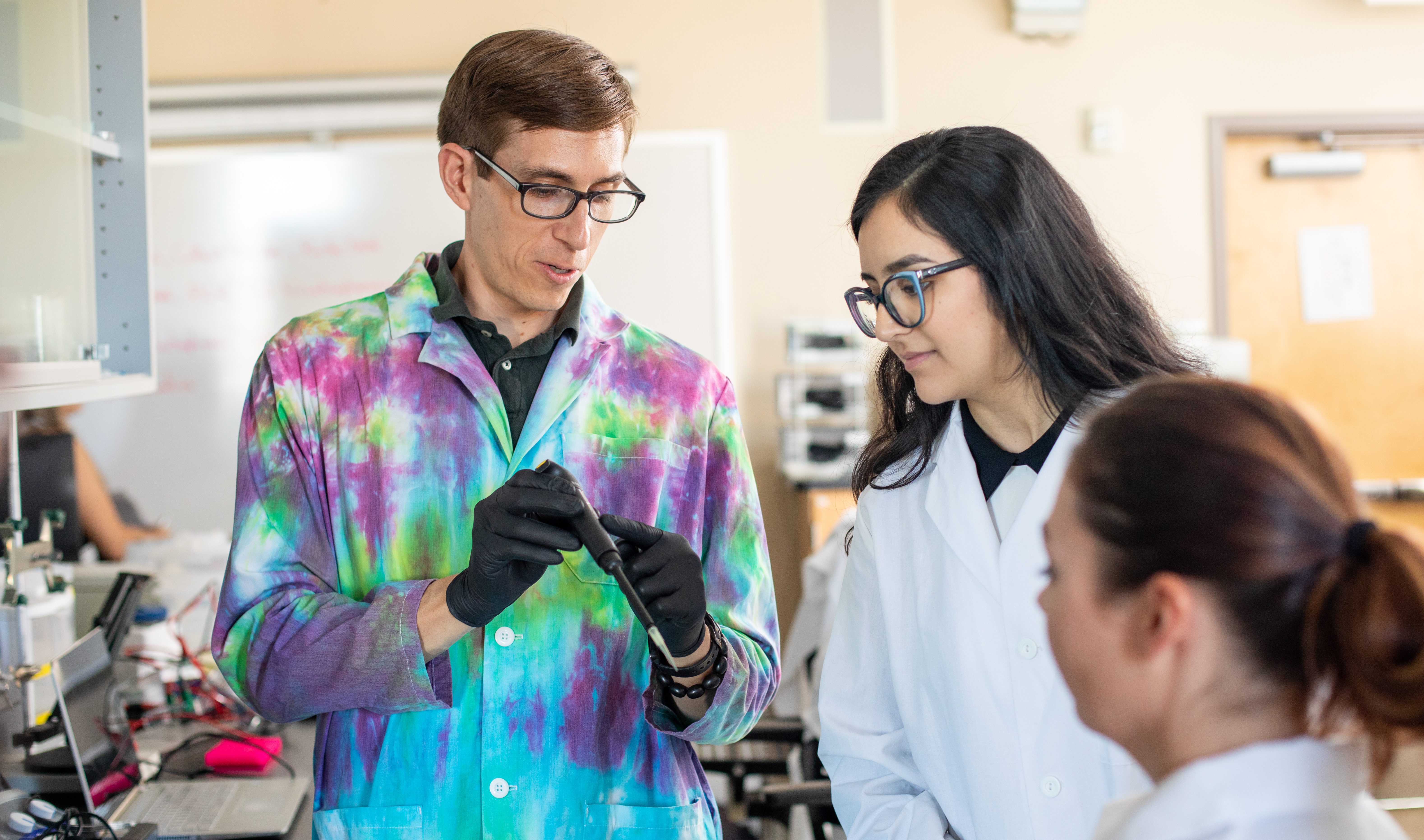 Un investigador de NAU en un laboratorio de ciencia le enseña una pipeta a dos estudiantes.