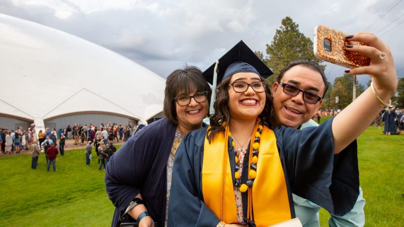 Female NAU graduate wearing cap, gown, and stole taking a selfie with family outside with the Skydome in the background