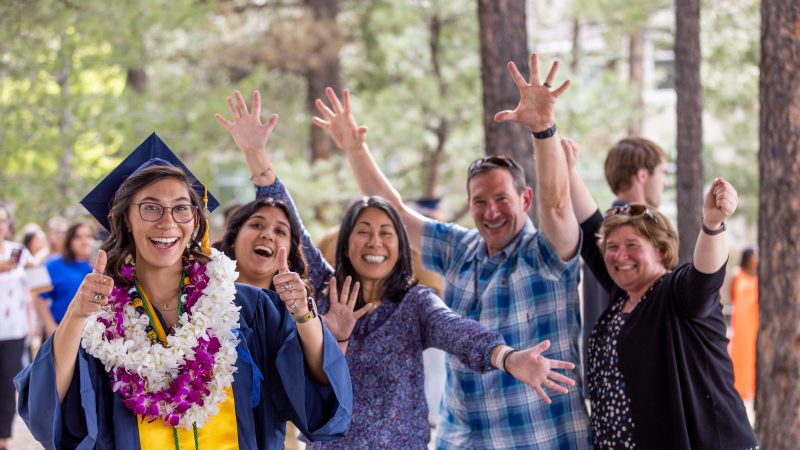 Female NAU graduate wearing cap, gown, stole, and leis giving thumbs up with her family outside