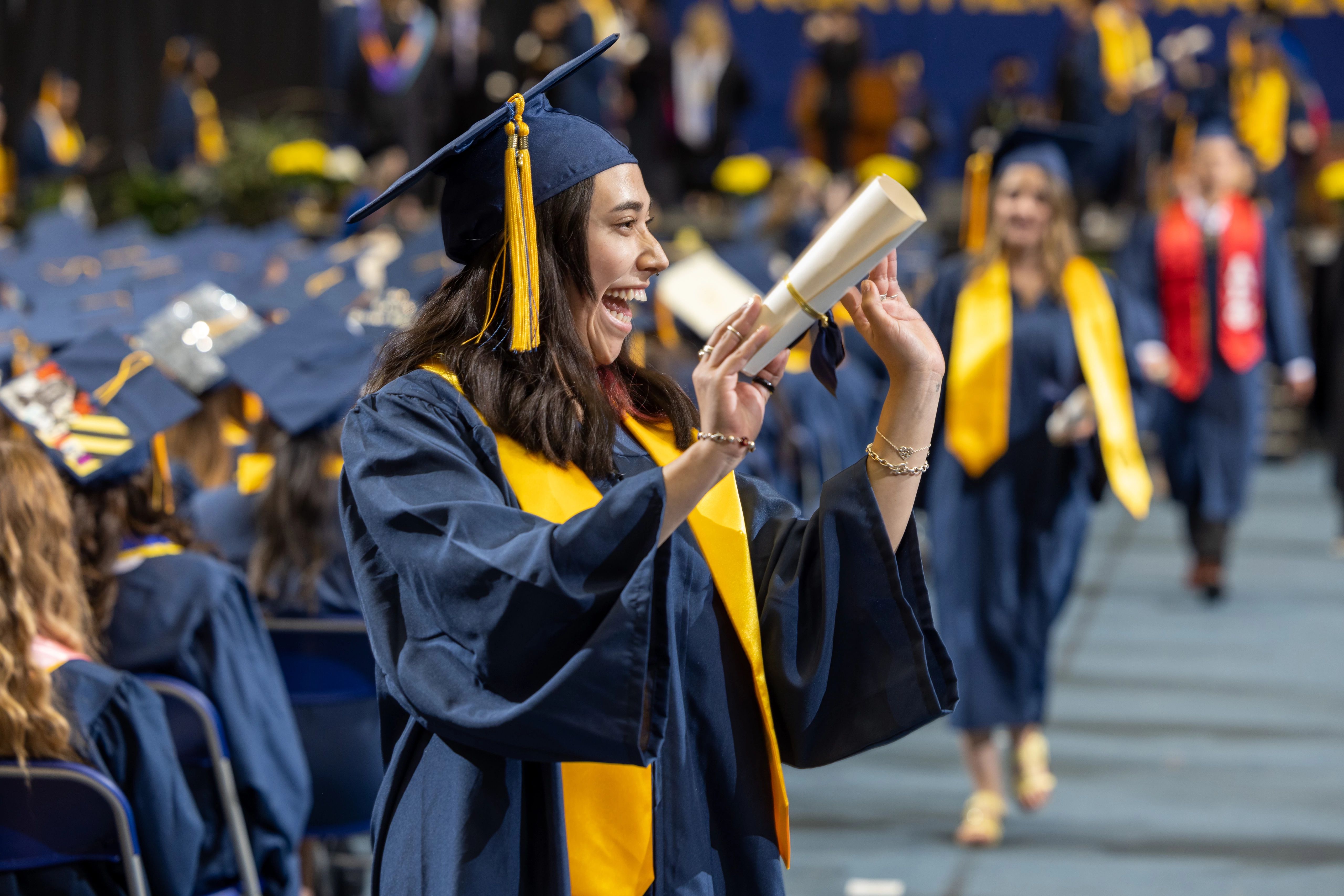 NAU grad walking down an aisle wearing regalia with diploma in hand waving to her family