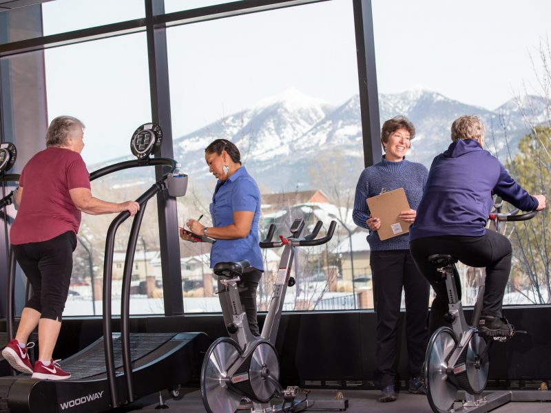 Two physical therapist speak with their patience on a stationary bike and a treadmill.