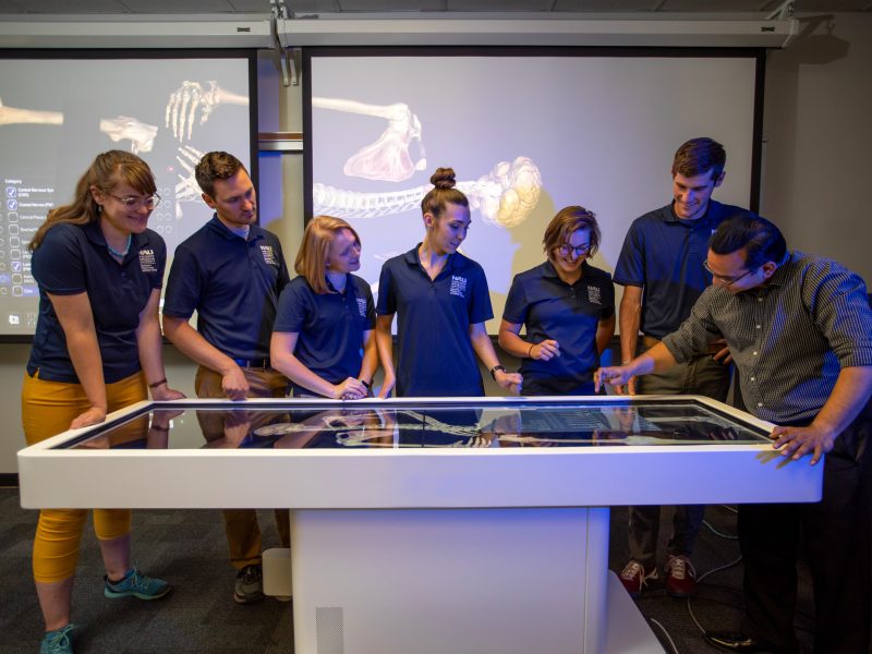 Students and a professor stand around a table observing a display of skeleton bones.