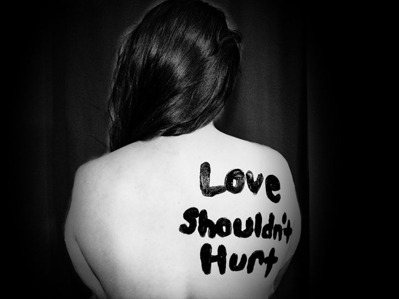 A black and white photograph of a woman's back, with the words, "Love Shouldn't Hurt" written on her shoulder.