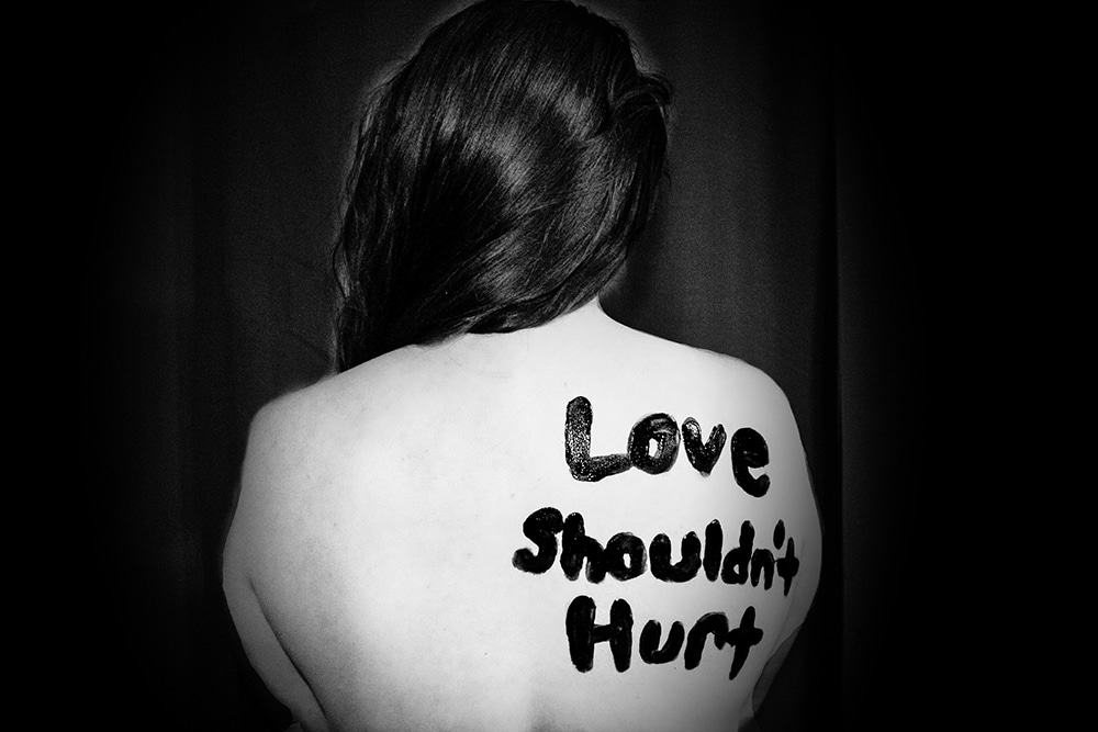 A black and white photograph of a woman's back, with the words, "Love Shouldn't Hurt" written on her shoulder.