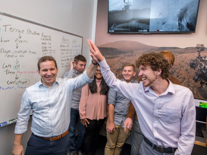 Students high fiving and smiling with a filled out whiteboard in the background.