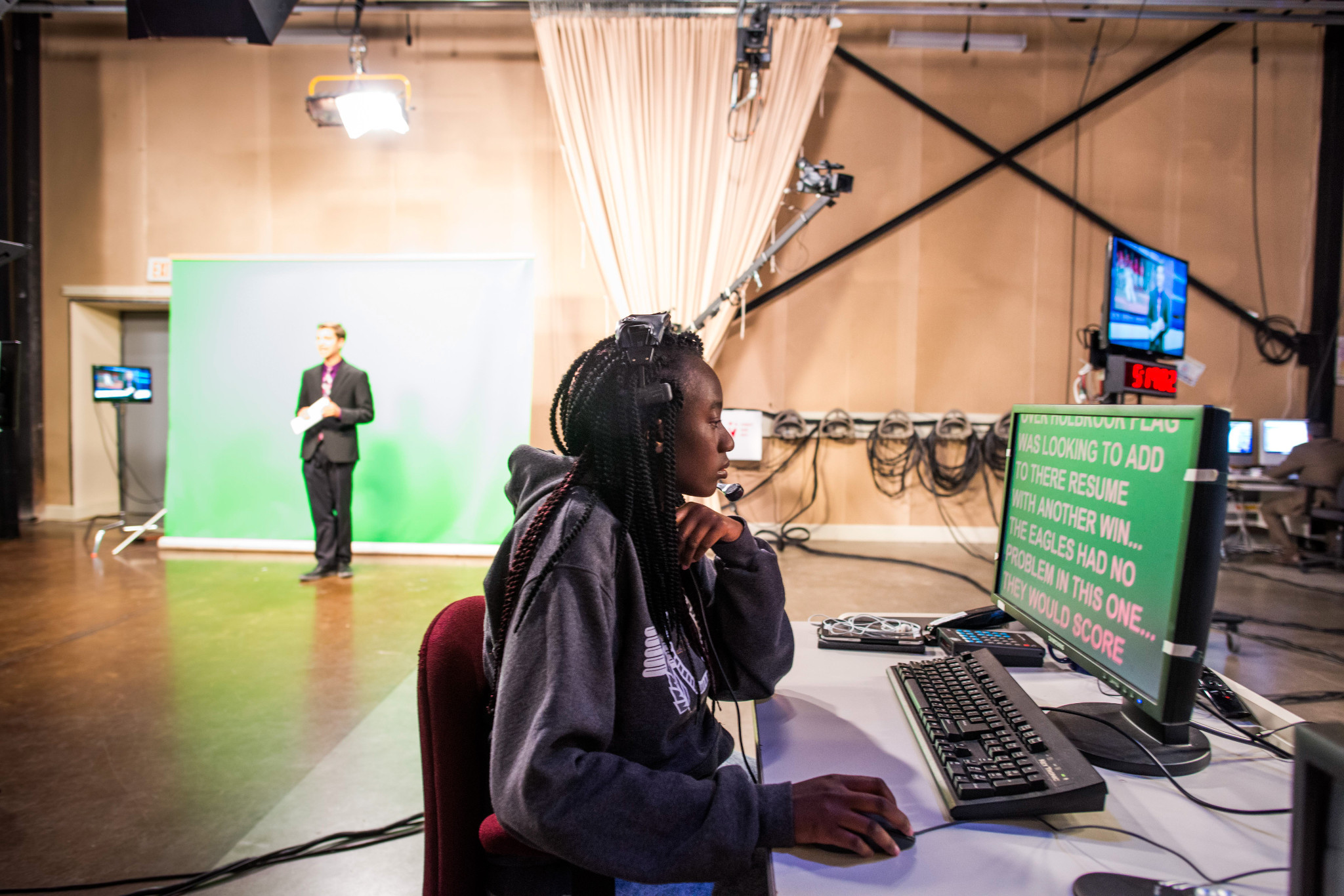 Student with a headset using a computer while TV production with another student infront of a green screen in the background.