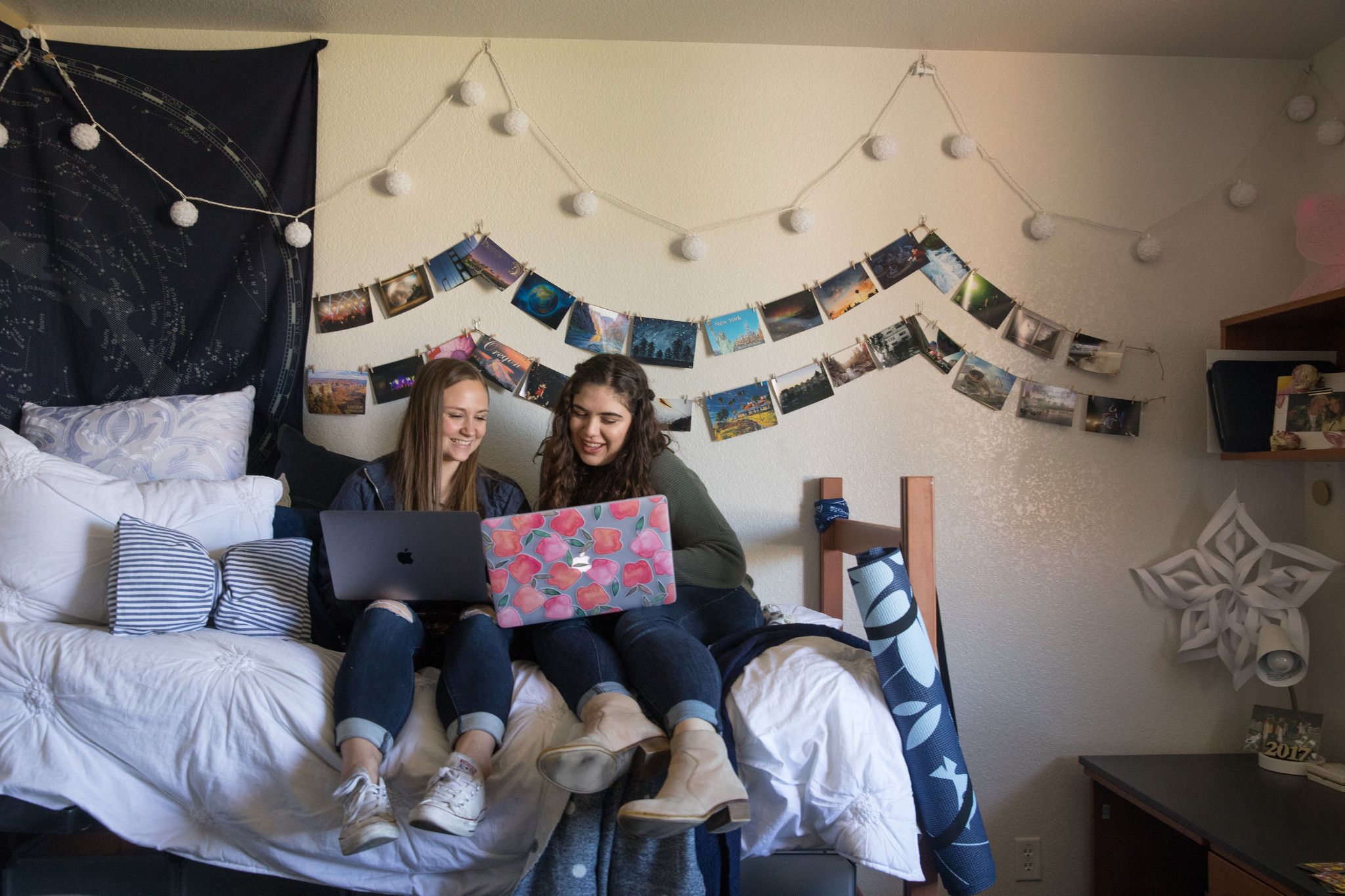 Two students sitting on a bed in a dorm while working on their laptops.