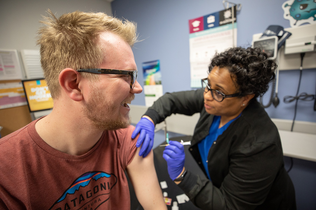 A student sits on a medical table as a nurse gives him a vaccine.