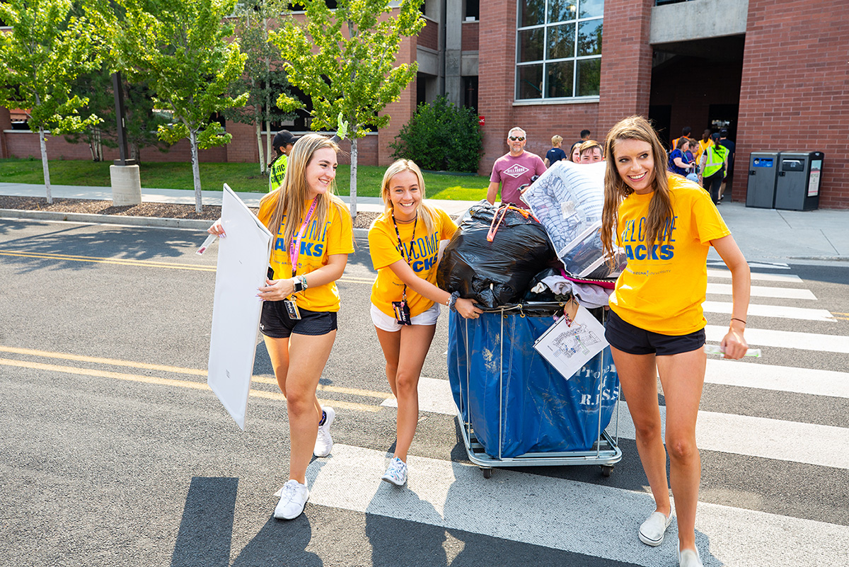 Volunteers in Welcome Jacks shirts help a new student push their blue cart full of luggage.