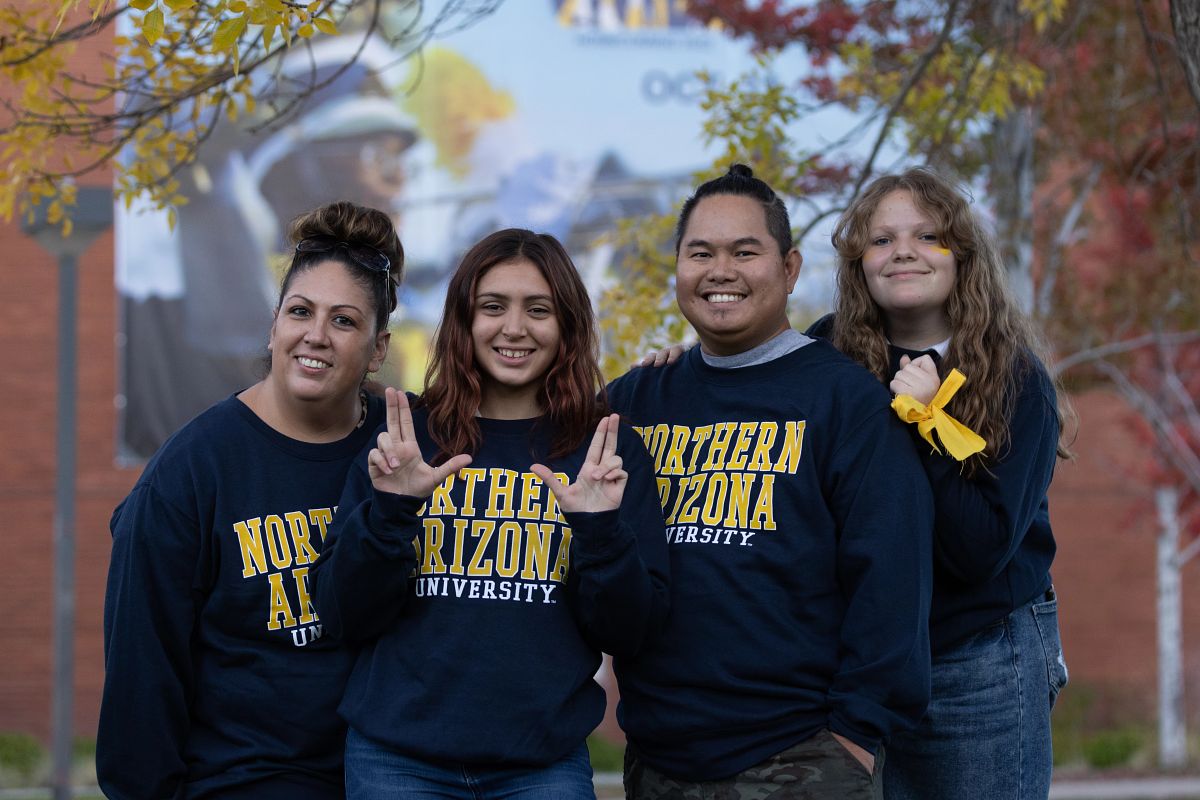 A family dressed in N A U sweatshirts smile at the camera and give the L J sign.