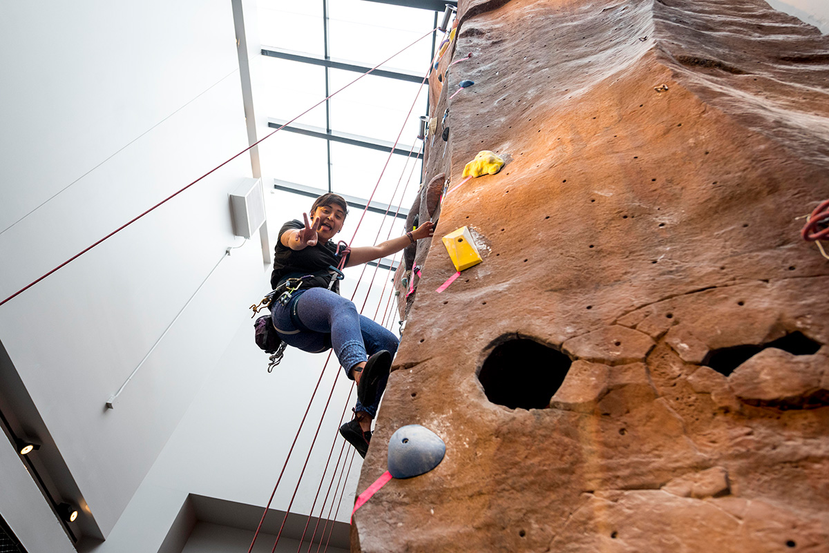 A student climbs the rock wall in campus rec. She looks down at the camera and gives a peace sign.