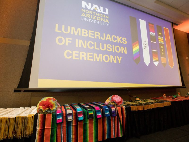 A series of colorful stoles sits on a table in front of a slide that reads "Lumberjacks of Inclusion."