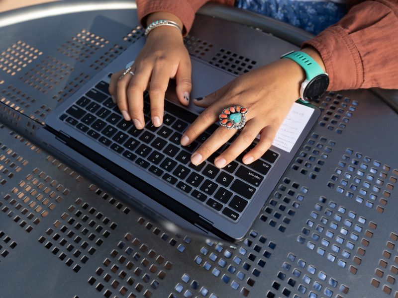 A student sits at a bench near the Native American Cultural Center and writes on their laptop.