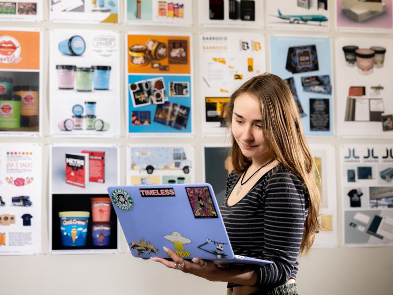 A Visual Communication student looks at her laptop and stands in front of a wall covered in graphic design print outs.