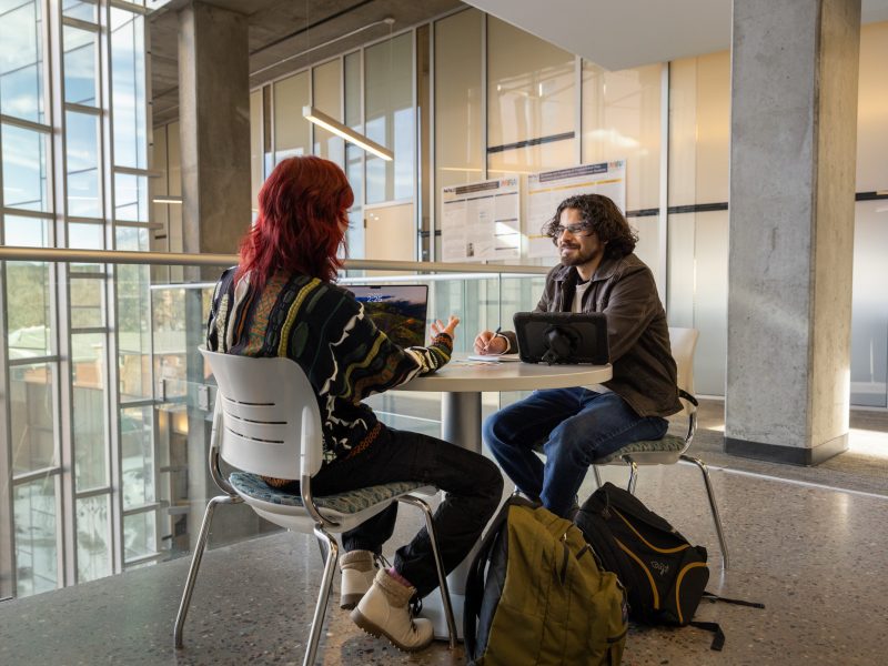 Students sit across from each other at a table inside a campus building.