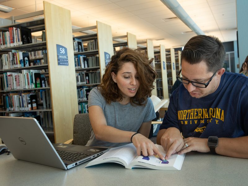 Two students hovering over the same book at a table in the library.