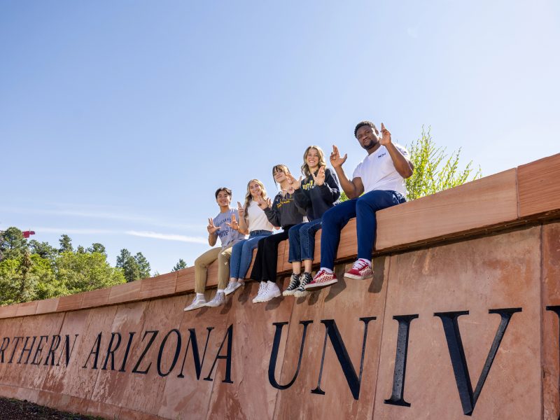 Students sitting on the N A U sign against a blue sky.