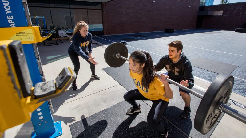 One female student holding a squat with a weight while two students spot.