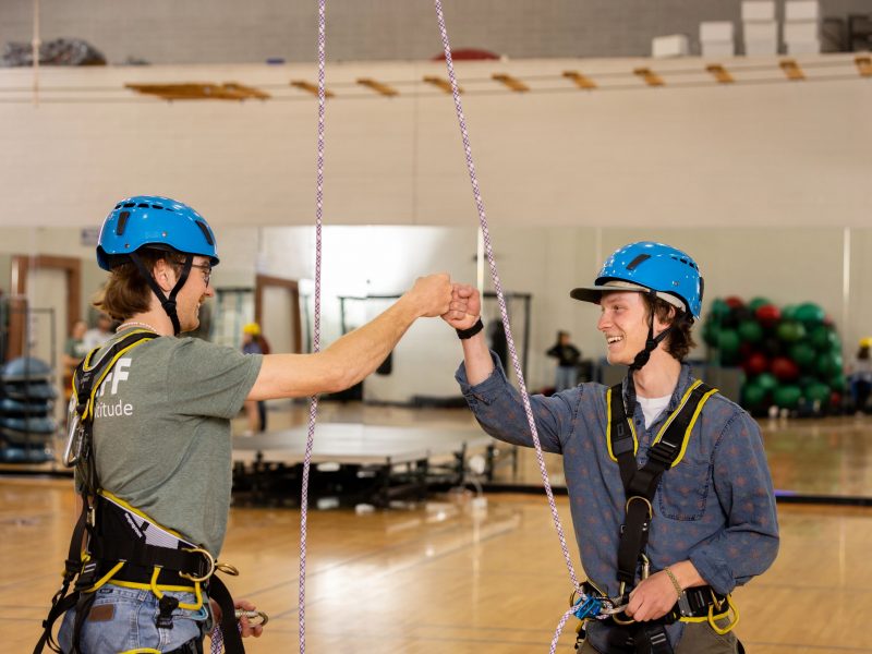 Two students fist bumping while attached to ropes on indoor rock climbing wall.