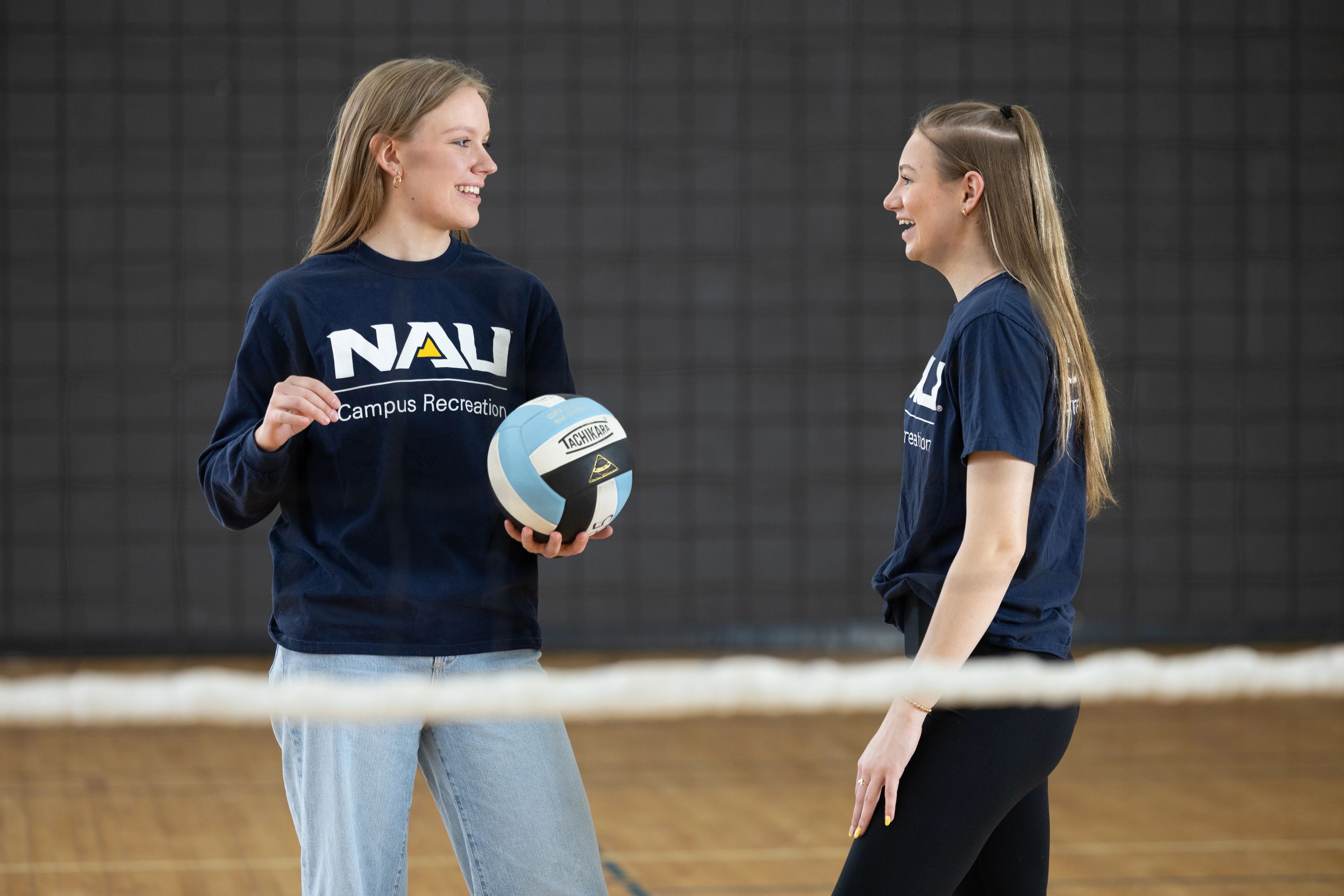 Female student holding a volleyball talking to another student in indoor volleyball court.