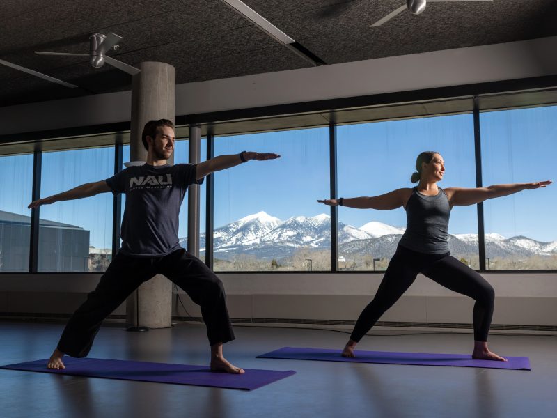 Two students performing standing yoga stretches with a smile, in front of a view of mountains from the window in the background.