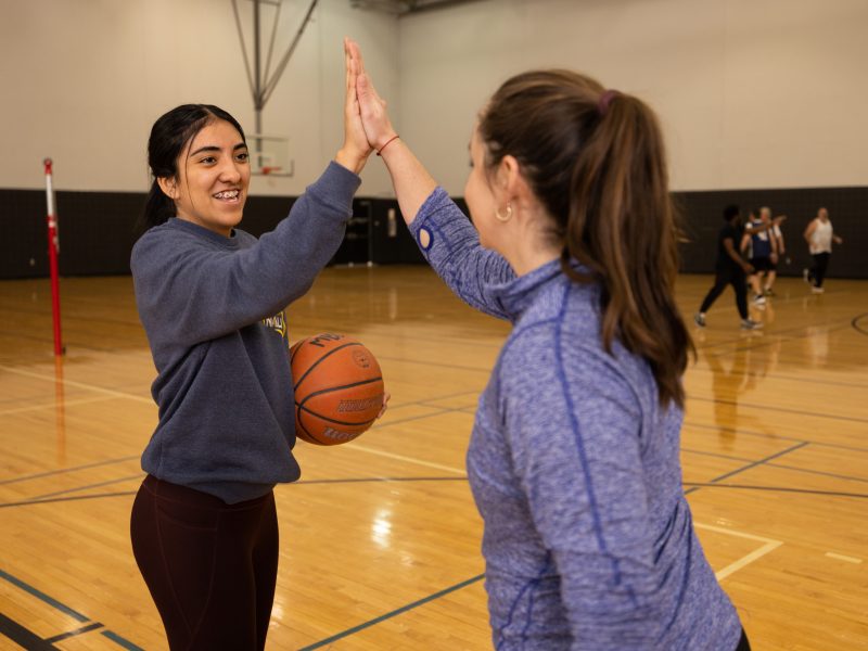 Two female student high five while one holds a basketball.