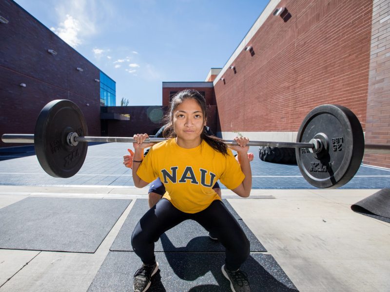 N A U student squatting outside holding barbell, being assisted by Campus Recreation trainers.