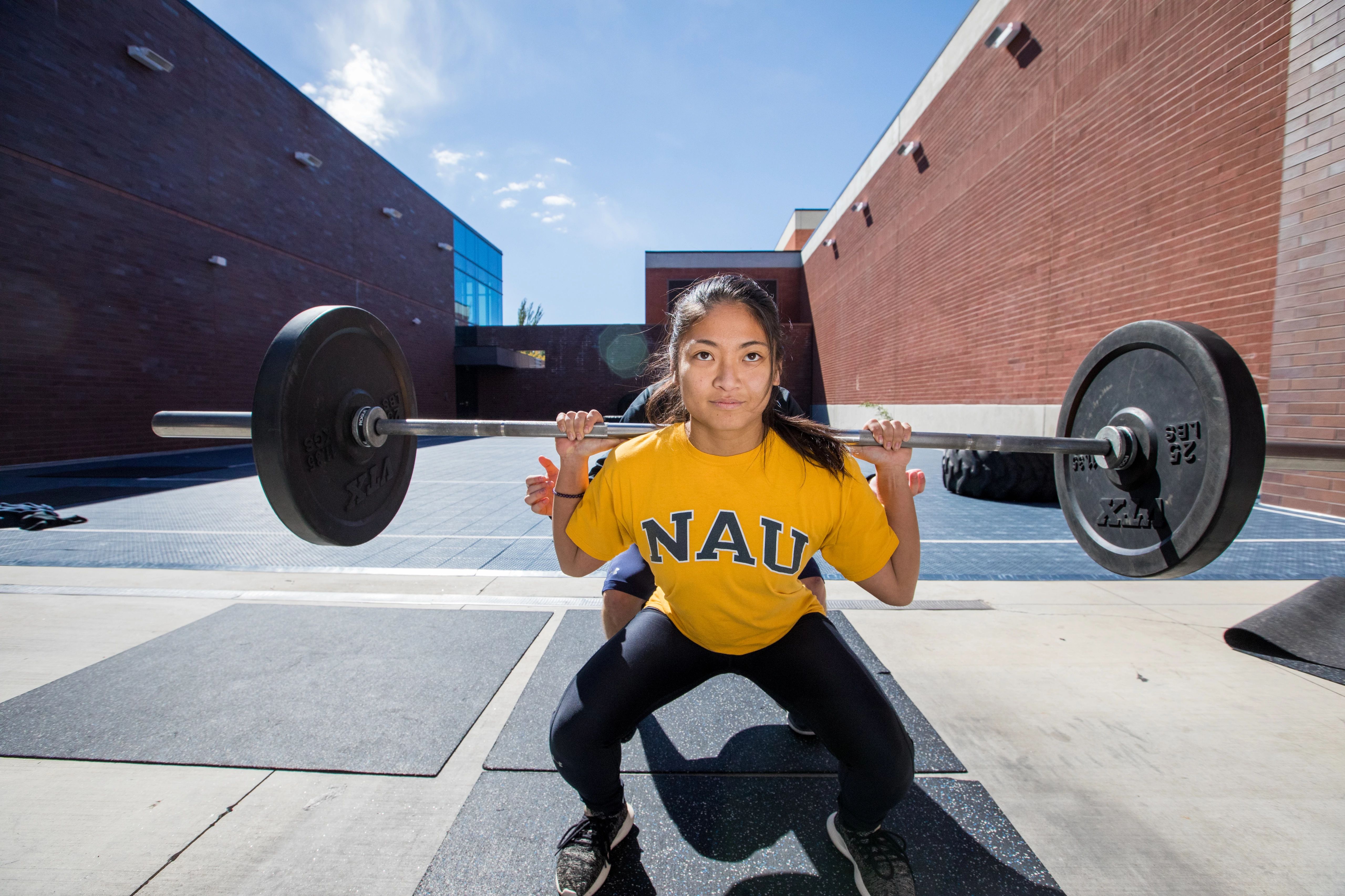 N A U student squatting outside holding barbell, being assisted by Campus Recreation trainers.