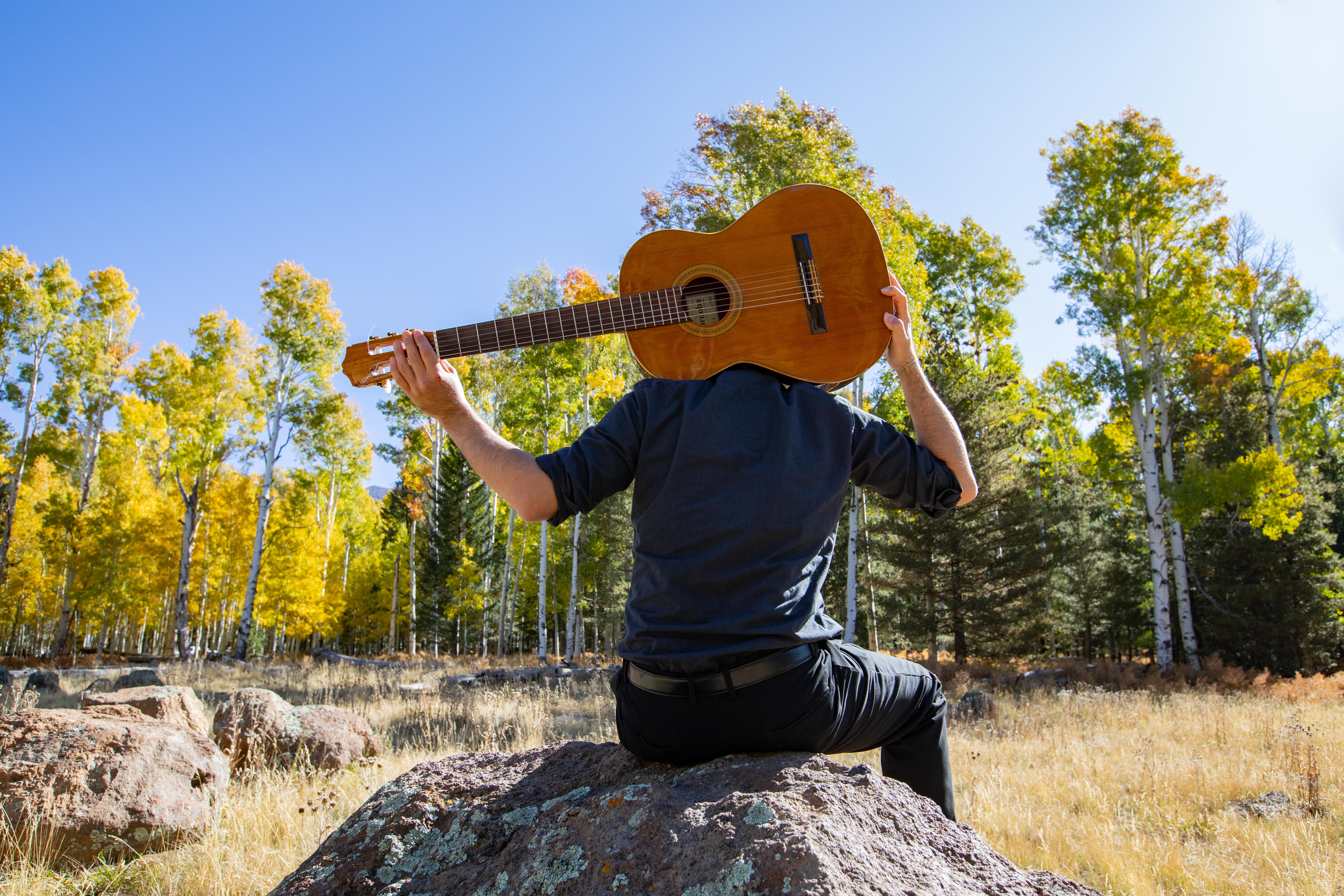 Person sitting on a rock in the forest holding a guitar behind their head.