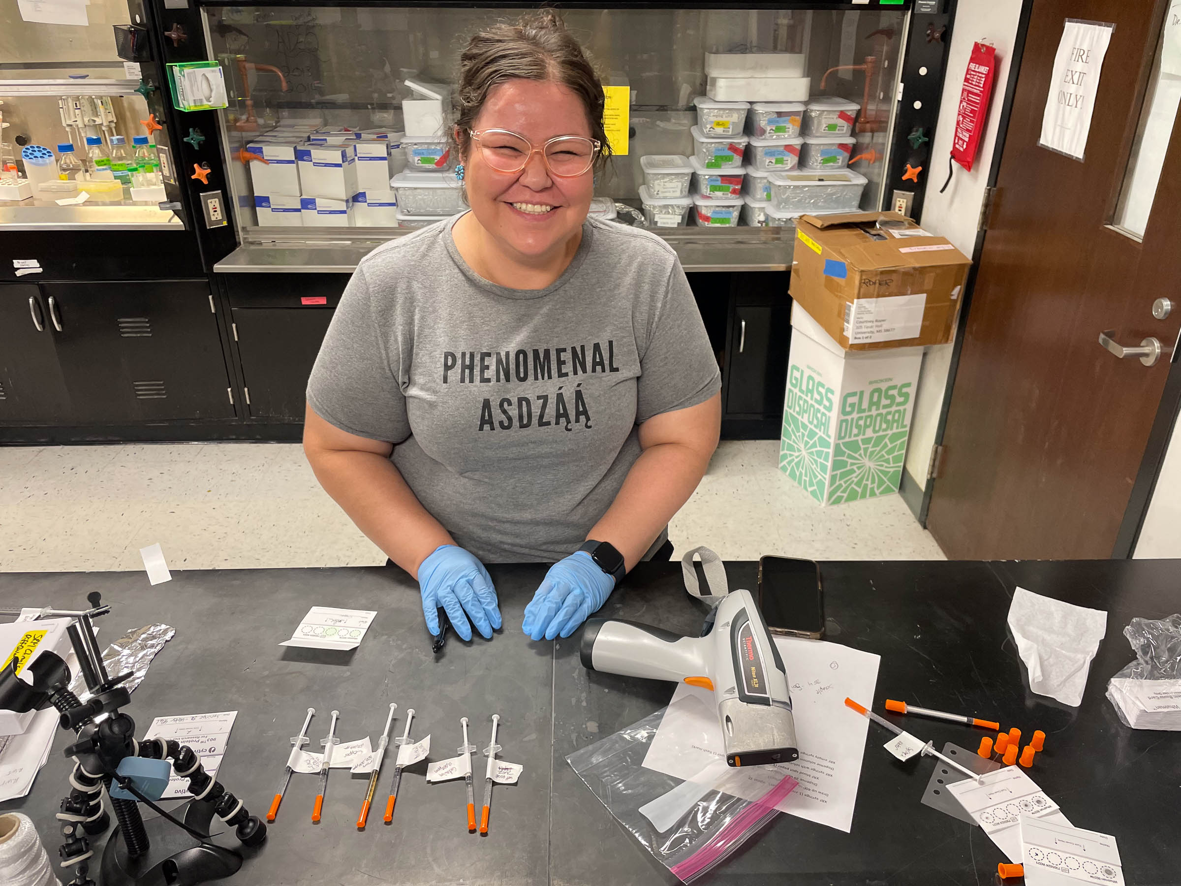 Goodluck prepares samples for analysis, wearing a shirt that says “phenomenal woman” in Diné.