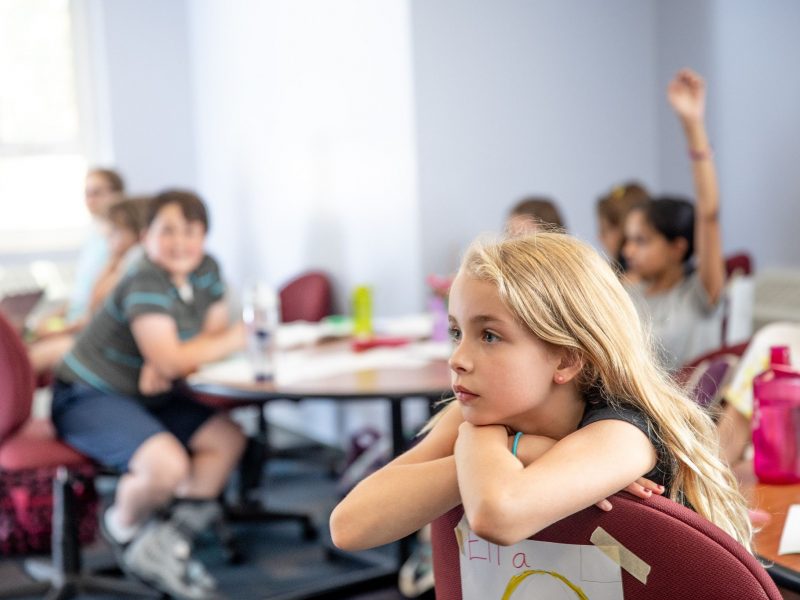 Child students sitting in classroom chairs listening to a teacher.