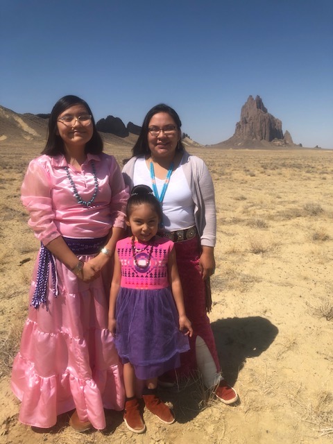 Two girls and their mother wear dresses while standing on reservation outdoor land.