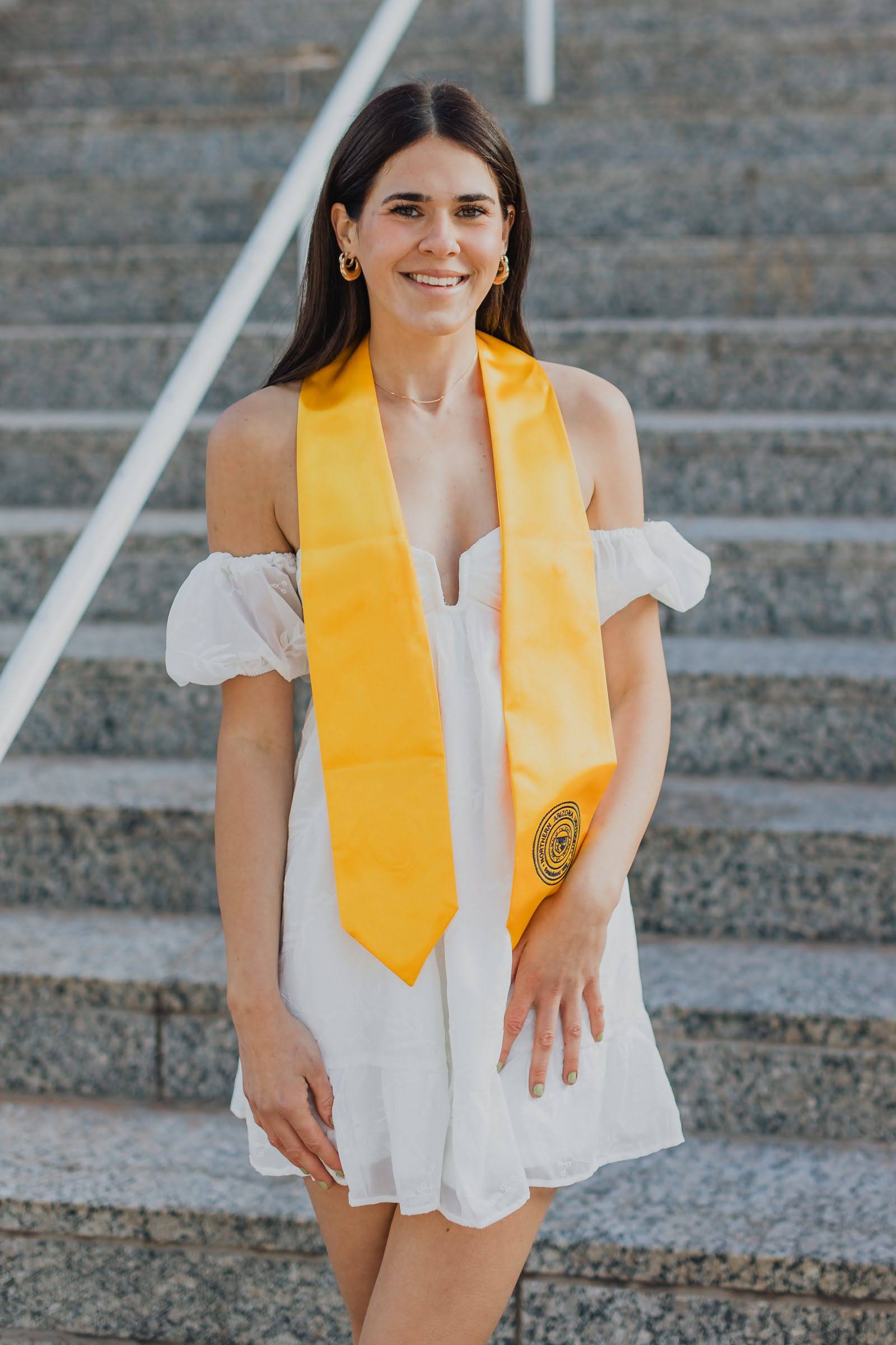 Alexis Henninger wearing a white dress with NAU graduation stole on the steps of the Babbitt building.