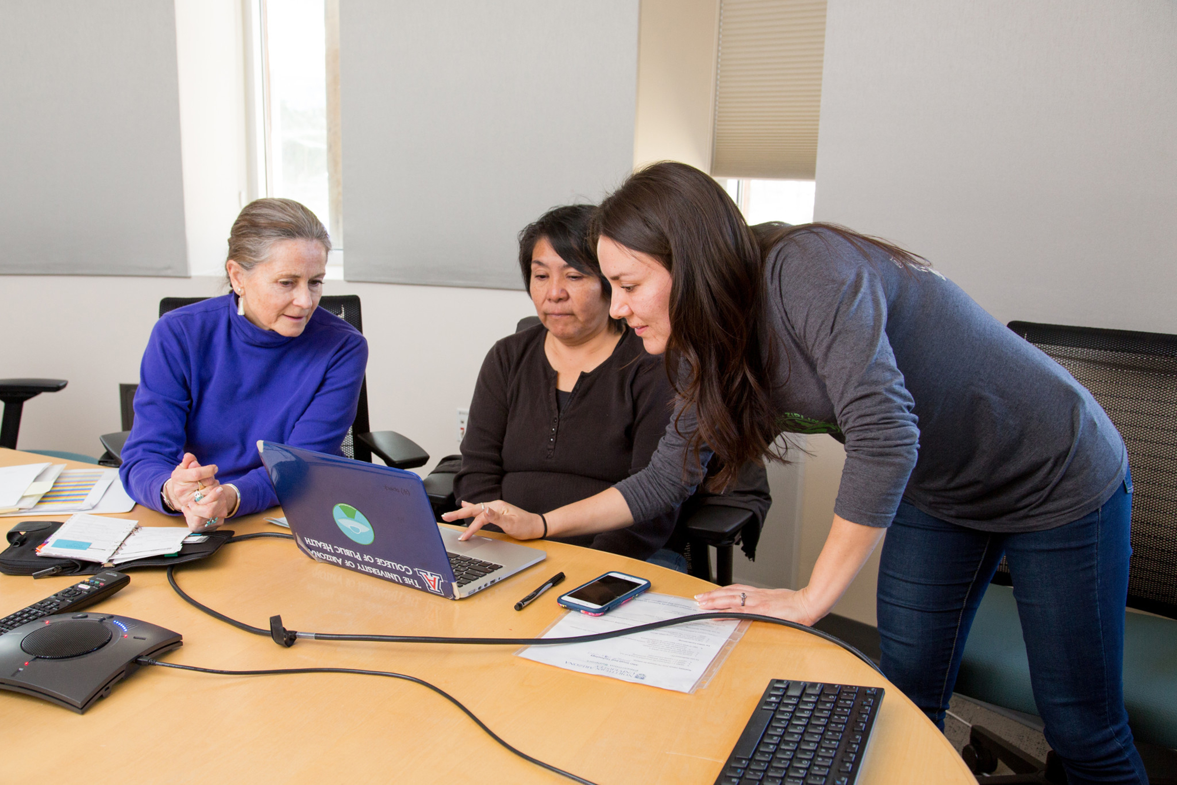 Amanda Hunter is leaning over her computer working with Nicolette Teufel-Shone and Bernita Paya on a report.