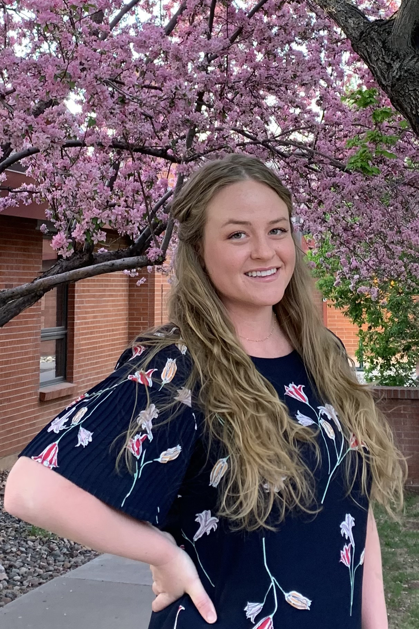 Amy Gelatt wearing a blue flowered dress standing by a blooming crabapple tree.