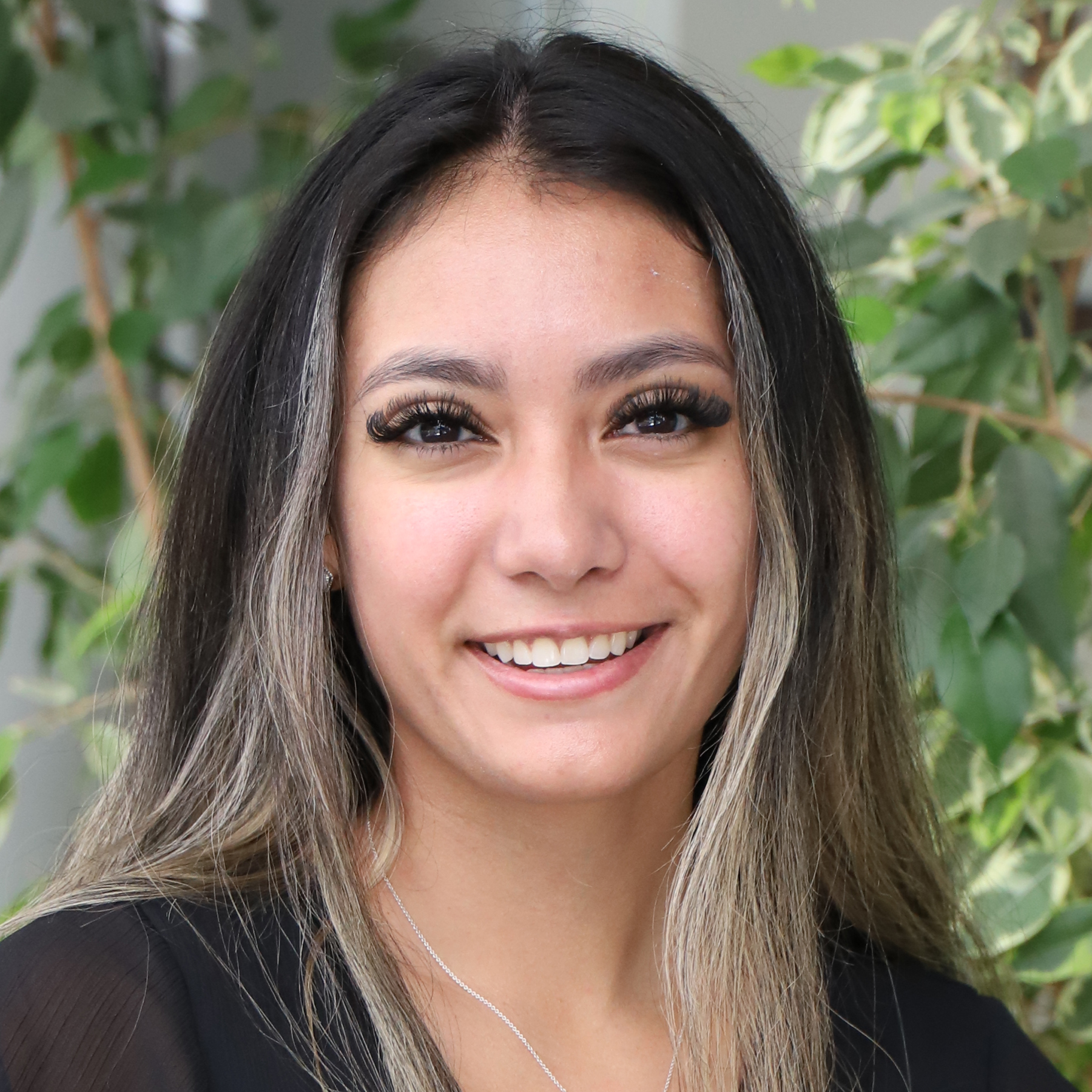 Angelica Alvarado is wearing a black top and with earrings and a necklace an standing in front of a plant.