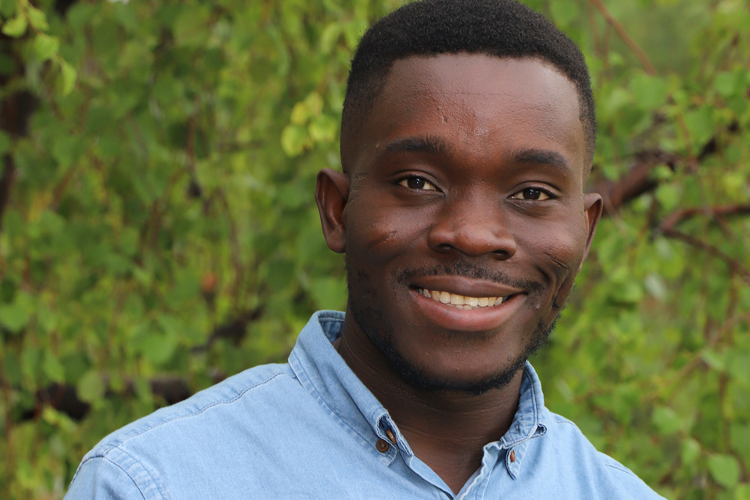 Kofi Mensah posing for a portrait outside in front of trees wearing a blue shirt.