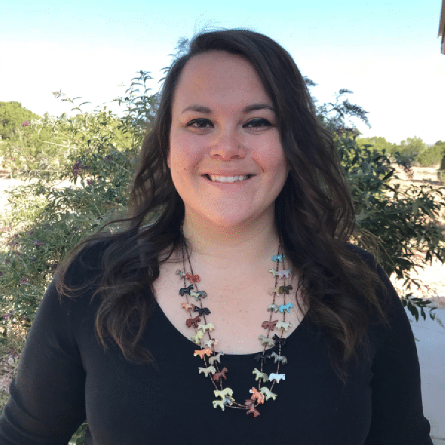 Carly Camplain is wearing a black top and Native American Jewelry and is standing in front of a large bush.