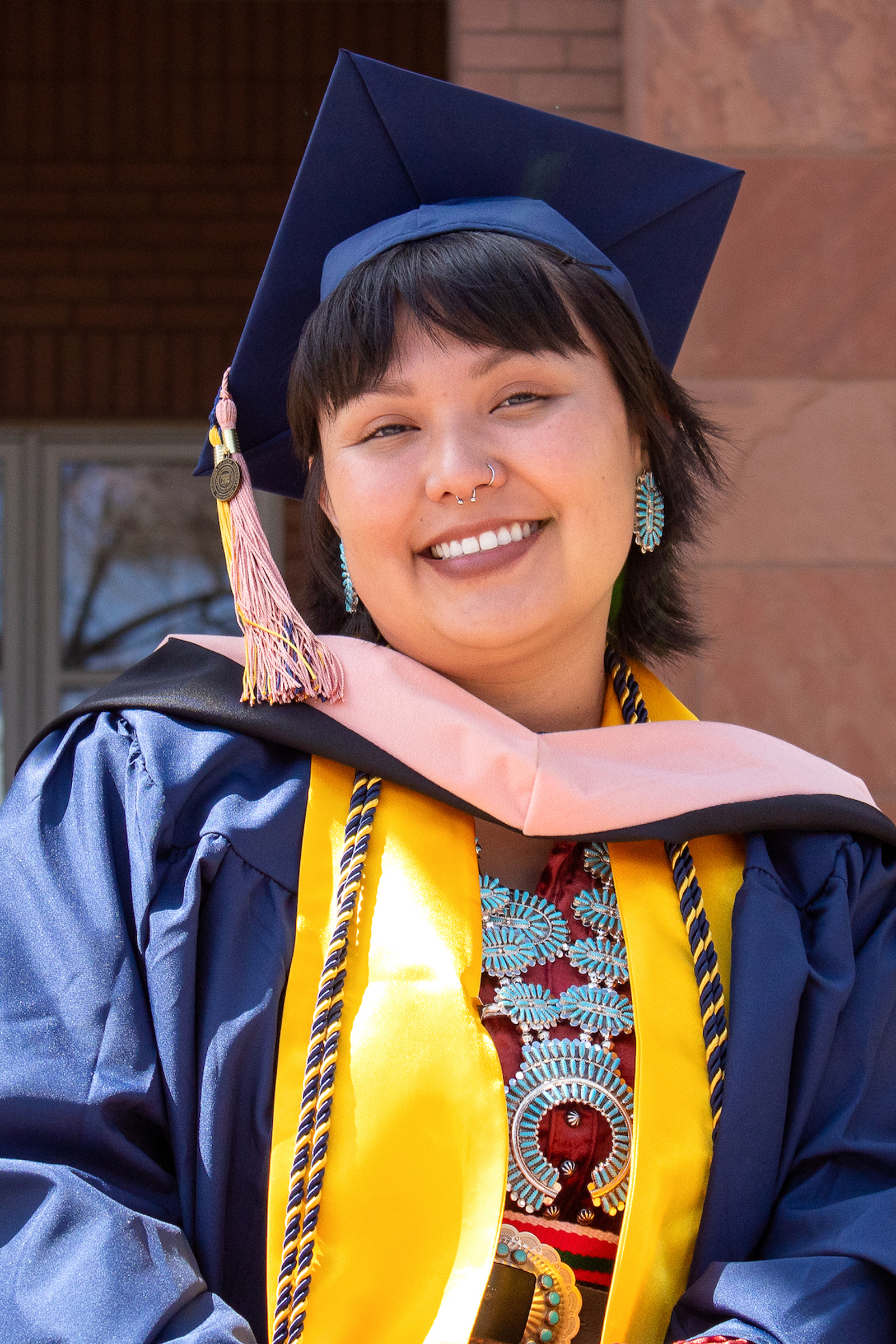 Chassity Begay standing by Cline Library wearing graduation gown, cap, stole, and Native American jewelry.