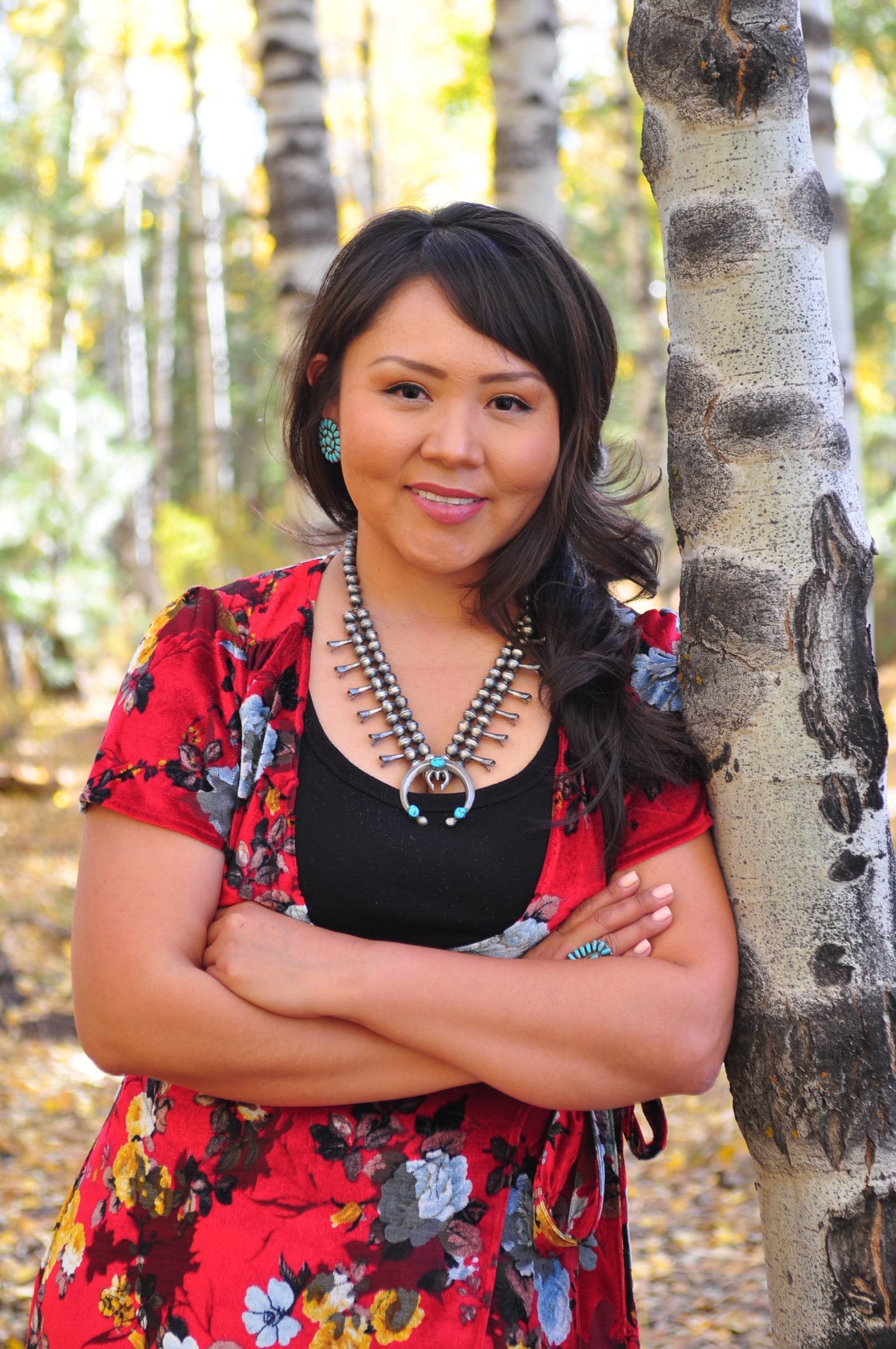 Jaime Begay is wearing a red floral and black top, with Native American jewelry, standing against a tree