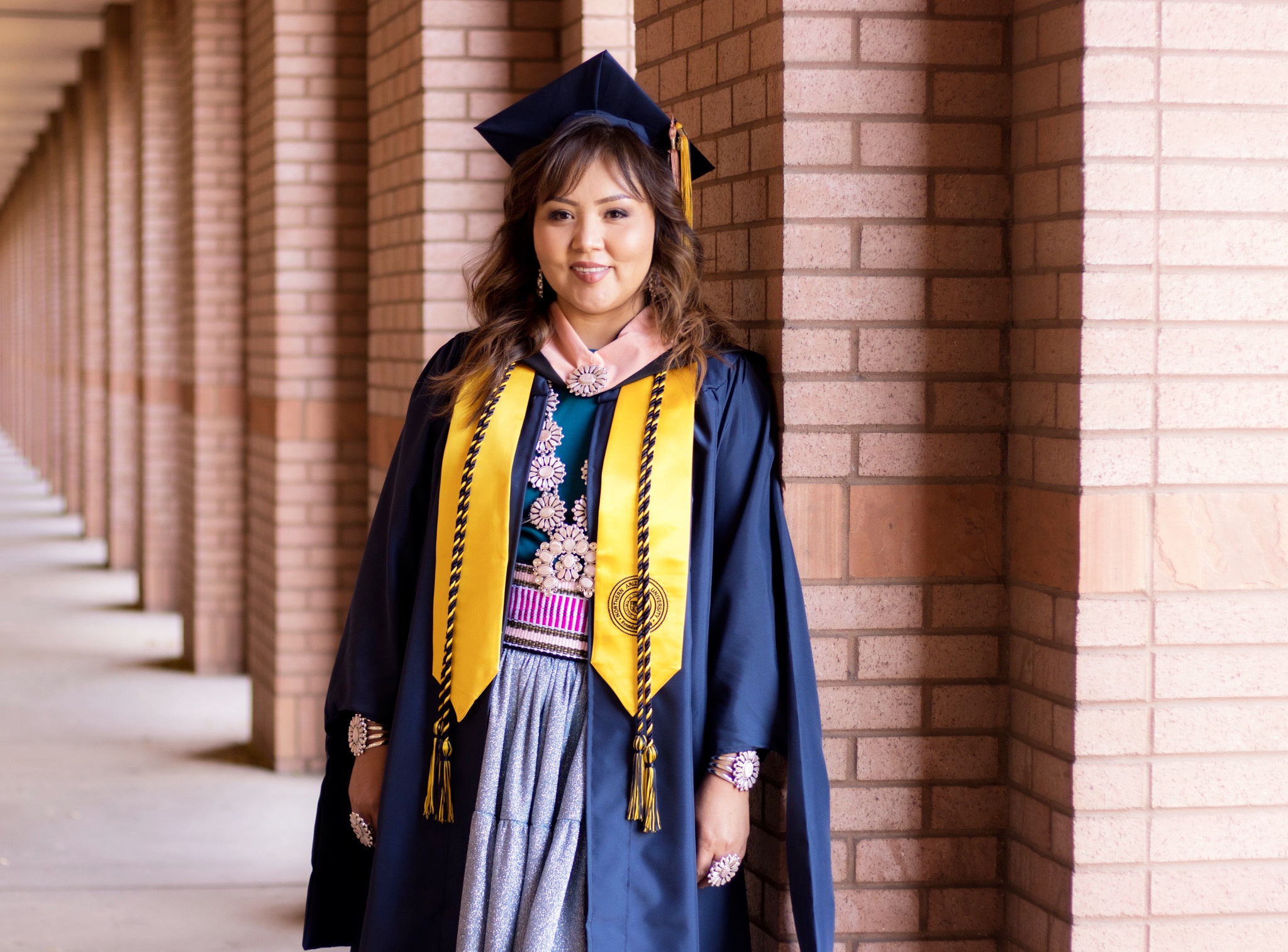 Jaime Begay standing outside the Cline Library wearing graduation own and cap.