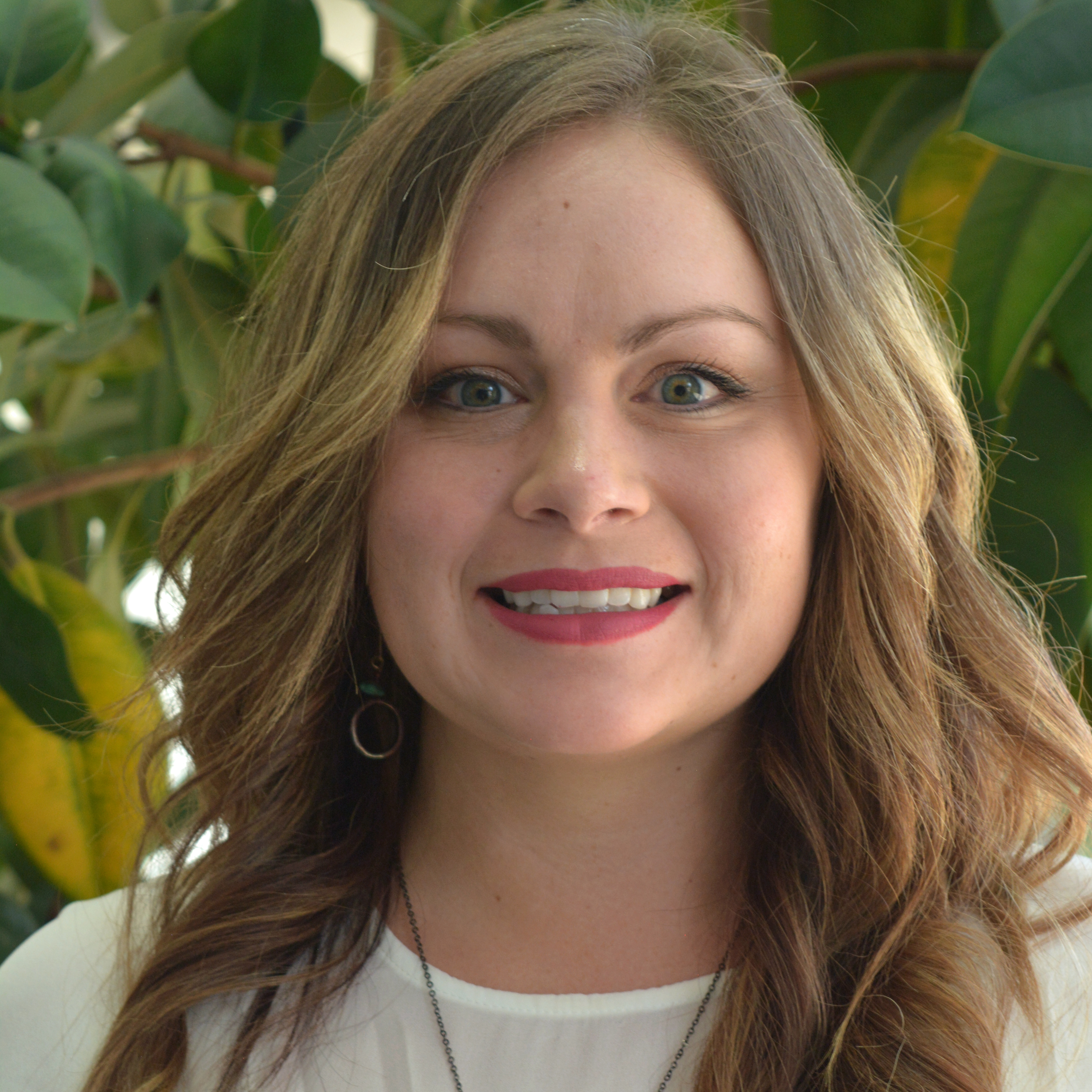 Melinda Smith is wearing a white top and necklace and earrings and is standing in front of a plant.