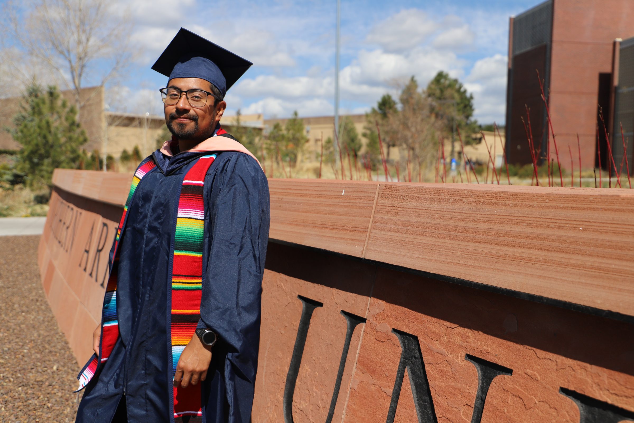 Omar Gomez in graduation attire next to N A U sign.